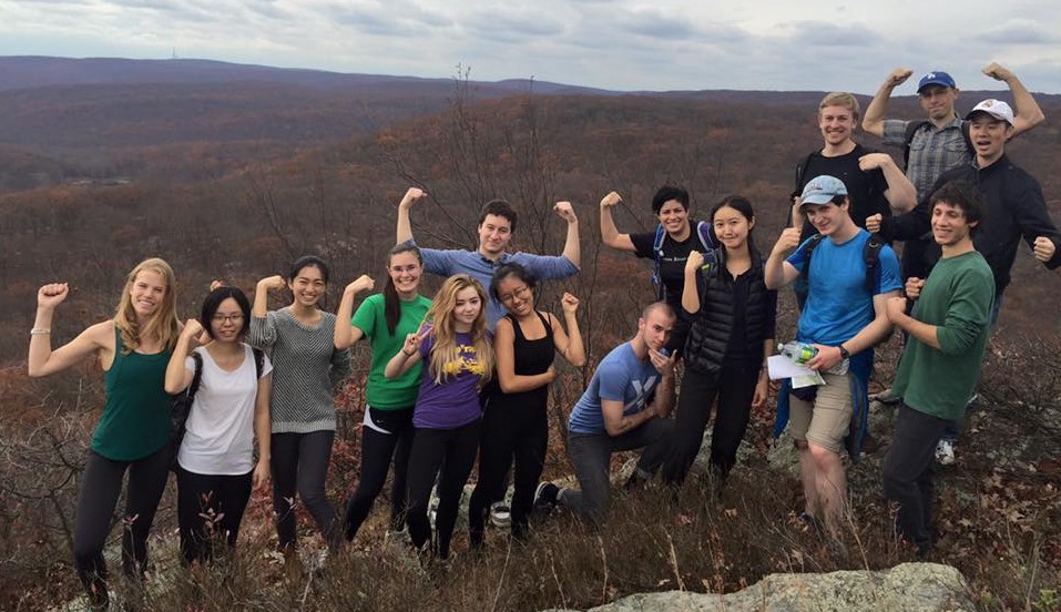 A group of fifteen students, members of Columbia's Hiking Club, pose in front of a scenic view on a cloudy day.